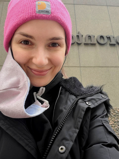 A selfie of Jodie, a white woman with brown hair and brown eyes in a black winter parka and pink Carhartt toque with a homemade mask hanging from one ear. Jodie is sitting in front of the Toronto Star headquarters with a peaceful and triumphant smile on her face.